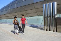 African-American woman and caucasian man with sports bag in hand walking together along wall of city construction — Photo de stock