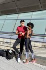 Personal trainer utilizzando smartphone e mostrando app per sorridere donna afroamericana mentre in piedi insieme accanto alla costruzione — Foto stock