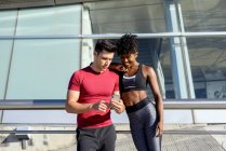 Personal trainer using smartphone and showing app to smiling African American woman while standing together next to building — Stock Photo
