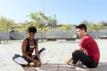 Africano mulher americana e homem caucasiano sentado e alongando-se para a frente enquanto pratica em conjunto na cidade no dia de verão — Fotografia de Stock