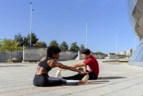 African American woman and caucasian man sitting and stretching forward while practicing together in city in summer day — Stock Photo