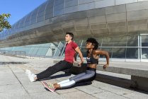 African American sportswoman and caucasian sportsman in red shirt doing push up exercises next to concrete bench in city — Stock Photo