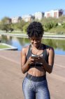Mujer afroamericana en el deporte que escucha música con teléfonos inteligentes y auriculares mientras entrena en la orilla del río en la ciudad. - foto de stock