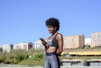African American woman in sportswear listening to music with smartphone and earphones while training at river bank in city — Stock Photo
