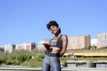 African American woman in sportswear listening to music with smartphone and earphones while training at river bank in city — Stock Photo