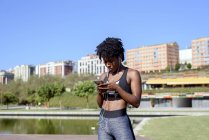 Donna afroamericana nello sportswear ascoltando musica con smartphone e auricolari mentre si allena in riva al fiume in città — Foto stock