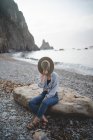 Turista escondido con sombrero disfrutando del paisaje marino mientras se enfría sobre piedra grande en la costa rocosa de Asturias mirando hacia otro lado - foto de stock