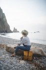 Turista femenina en sombrero disfrutando del paisaje marino mientras se enfría sobre una gran piedra en la costa rocosa de Asturias mirando hacia otro lado - foto de stock