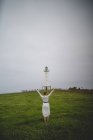 Vue arrière de la femme en robe blanche marchant prudemment dans le champ contre le phare par temps nuageux dans les Asturies, Espagne — Photo de stock