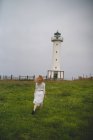 Back view of woman in white dress walking thoughtfully in field against lighthouse in cloudy weather in Asturias, Spain — Stock Photo