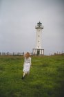 Vue arrière de la femme en robe blanche marchant prudemment dans le champ contre le phare par temps nuageux dans les Asturies, Espagne — Photo de stock