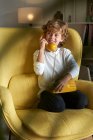 Niño con ropa casual sentado en un cómodo sillón amarillo y jugando a hablar por teléfono retro - foto de stock