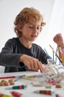 Little boy with curly hair smiling while sitting at table and painting against white wall at home — Stock Photo