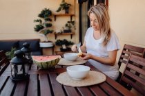 Pregnant woman in white home clothes cutting banana to white bowl at terrace — Stock Photo
