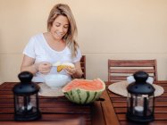 Pregnant woman in white home clothes cutting banana to white bowl at terrace — Stock Photo