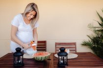 Schwangere in weißer Hauskleidung schneidet Wassermelone in weiße Schüssel auf der Terrasse — Stockfoto