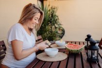 Side view of pregnant woman in white home clothes cutting banana to white bowl at terrace — Stock Photo