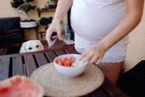 Abgeschnittenes Bild einer schwangeren Frau, die Wassermelone auf der Terrasse schneidet, während Hund hinter Holztisch sitzt und zusieht — Stockfoto