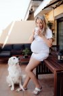 Zufriedene schwangere Frau in weißem T-Shirt und kurzer Hose, die morgens auf der Terrasse Kaffee trinkt, während Labrador-Hund neben ihr sitzt — Stockfoto