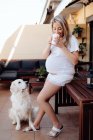 Mujer embarazada con camiseta blanca y pantalones cortos bebiendo café en la terraza en la mañana con perro labrador - foto de stock