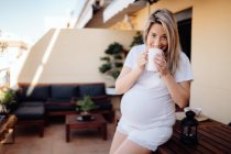 Alegre mujer embarazada rubia apoyada en la mesa de madera en la terraza mientras bebe té de la mañana y mira a la cámara - foto de stock