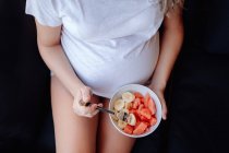 Imagen recortada de una mujer embarazada comiendo plátanos en rodajas y sandía de un tazón con tenedor mientras está sentada en un sofá oscuro - foto de stock