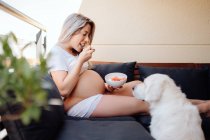 Весела блондинка вагітна жінка в білому домашньому одязі годує лабрадора собакою з шматком банана з чаші — стокове фото