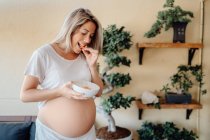 Zufriedene blonde schwangere Frau, die zu Hause mit Pflanzen gegen Wand steht und nackten Bauch berührt, während sie Schüssel in der Hand hält — Stockfoto