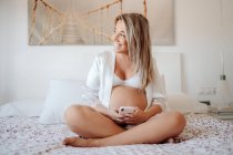 Content blonde pregnant woman in white open shirt and bra browsing mobile phone while sitting with crossed legs on big bed in bright room — Stock Photo