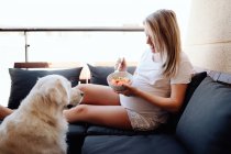 Весела блондинка вагітна жінка в білому домашньому одязі годує лабрадора собакою з шматком банана з миски, сидячи на дивані на терасі — стокове фото
