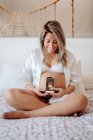 Беременная женщина демонстрирует изображение ультразвукового сканирования на смартфоне, сидя в лифчике и открытой рубашке на кровати со скрещенными ногами — стоковое фото