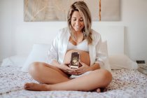 Mujer embarazada demostrando imagen de ultrasonido escaneado en el teléfono inteligente mientras está sentado en sujetador y camisa abierta en la cama con las piernas cruzadas - foto de stock
