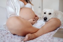 Image recadrée de la femme enceinte assise sur le lit avec les jambes croisées et toucher le ventre — Photo de stock