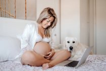 Sonriente mujer embarazada rubia sentada en la cama con las piernas cruzadas y tocando el vientre - foto de stock