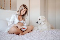 Content blonde pregnant woman sitting on bed with crossed legs holding bowl of food while Labrador dog putting paws near and looking at meal — Stock Photo