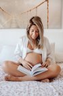 Content blonde pregnant woman wearing white elegant home clothes reading book while sitting with crossed legs on bed in bright room — Stock Photo