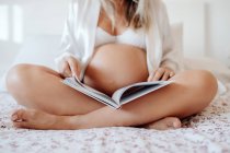 Zugeschnittenes Bild einer schwangeren Frau in weißer Hauskleidung, die Buch liest, während sie mit überkreuzten Beinen auf dem Bett im hellen Zimmer sitzt — Stockfoto