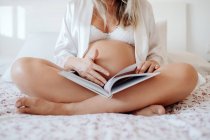 Cropped image of pregnant woman in white home clothes reading book while sitting with crossed legs on bed in bright room — Stock Photo