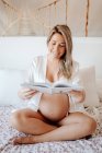 Content blonde pregnant woman in home clothes reading book while sitting with crossed legs on bed in bright room — Stock Photo