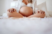 Abgeschnittenes Bild einer schwangeren Frau in weißer offener Bluse und BH, die mit überkreuzten Beinen auf dem Bett sitzt — Stockfoto