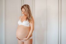 Happy blonde pregnant woman dressed in white bra and panties holding belly while standing against bright wall — Stock Photo