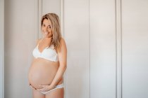 Mulher grávida loira feliz vestida com sutiã branco e calcinha segurando barriga enquanto está de pé contra a parede brilhante e olhando para a câmera — Fotografia de Stock