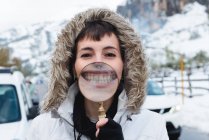 Женщина с пирсингом носа в белой зимней куртке с капюшоном на голове смотрит в камеру улыбающиеся зубы по лупе — стоковое фото