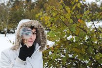 Woman with nose piercing wearing white winter jacket with hood on head looking at camera through magnifying glass — Stock Photo