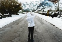 Rückansicht einer Person in weißer Winterjacke mit Kapuze auf dem Kopf, die mitten auf einer asphaltierten Straße am Fuße der Berge bei verschneitem Wetter steht — Stockfoto