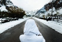 Visão traseira de pessoa em jaqueta de inverno branca com capuz na cabeça em pé no meio da estrada de asfalto a pé de montanhas em tempo nevado — Fotografia de Stock