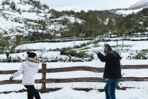 Hombre y mujer gozosos usando ropa de invierno jugando alegremente con bolas de nieve mientras están de vacaciones en el pueblo de montaña - foto de stock