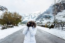 Mujer en chaqueta blanca de invierno con capucha y pantalones negros tomando fotos con la cámara mientras está de pie en medio de la carretera de asfalto - foto de stock