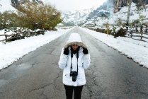 Woman in white winter jacket with hood and black pants taking picture with camera while standing in middle of asphalt road — Stock Photo