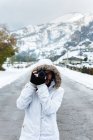 Femme en veste d'hiver blanche avec capuche et pantalon noir prenant des photos avec caméra tout en se tenant au milieu de la route asphaltée — Photo de stock